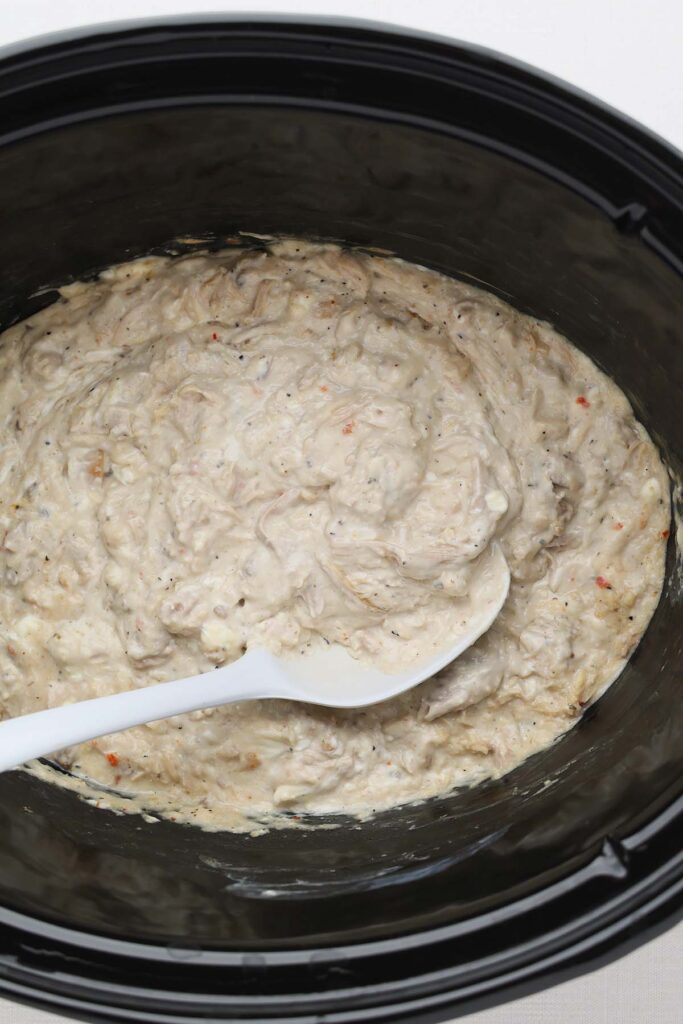 Creamy shredded chicken mixture in a slow cooker with a white spoon, ready to be served over pasta.