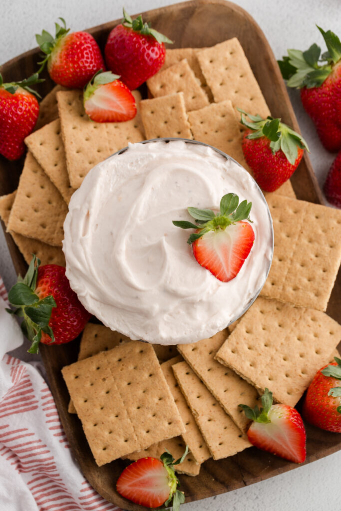 Top down image showing a bowl of pink dip with a strawberry that has been sliced in half on top. The bowl is located on a tray with graham crackers and more berries
