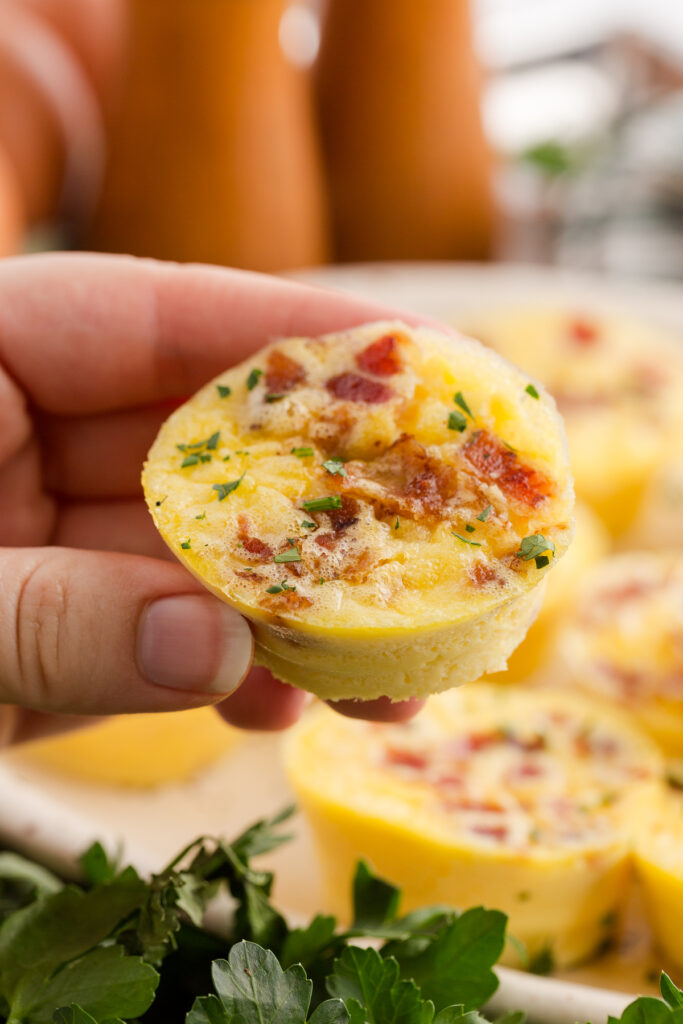 close up image showing a hand holding a single egg bite that has bacon and chives on top