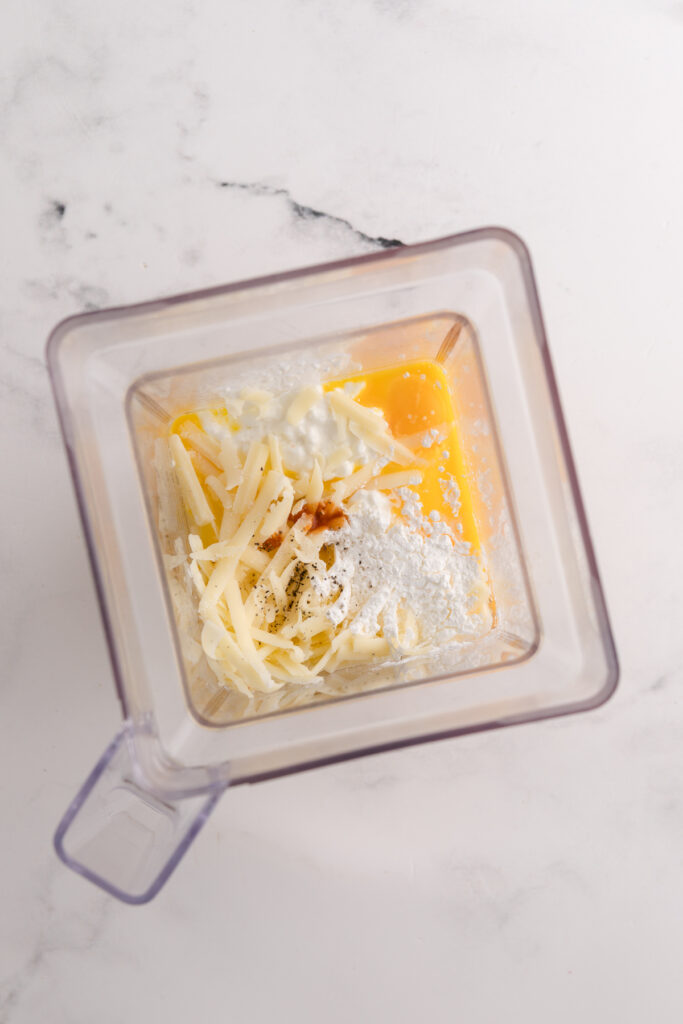 top down image showing the inside of a blender with cheese, eggs, cottage cheese, ranch seasoning unblended