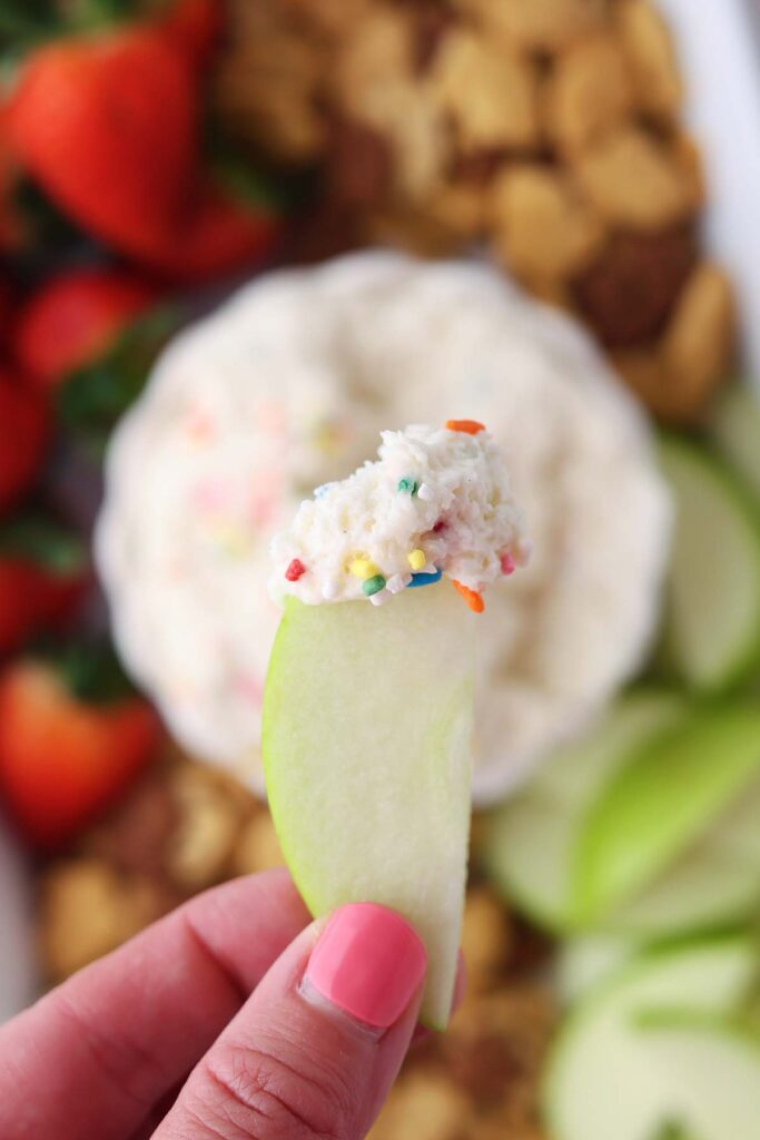 Creamy birthday cake dip topped with rainbow sprinkles in a white bowl, surrounded by fresh strawberries, green apple slices, and assorted animal crackers on a dessert boa