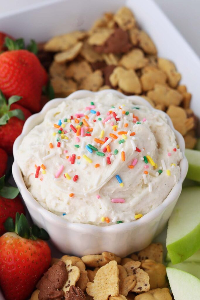 Creamy birthday cake dip topped with rainbow sprinkles in a white bowl, surrounded by fresh strawberries, green apple slices, and assorted animal crackers on a dessert boa