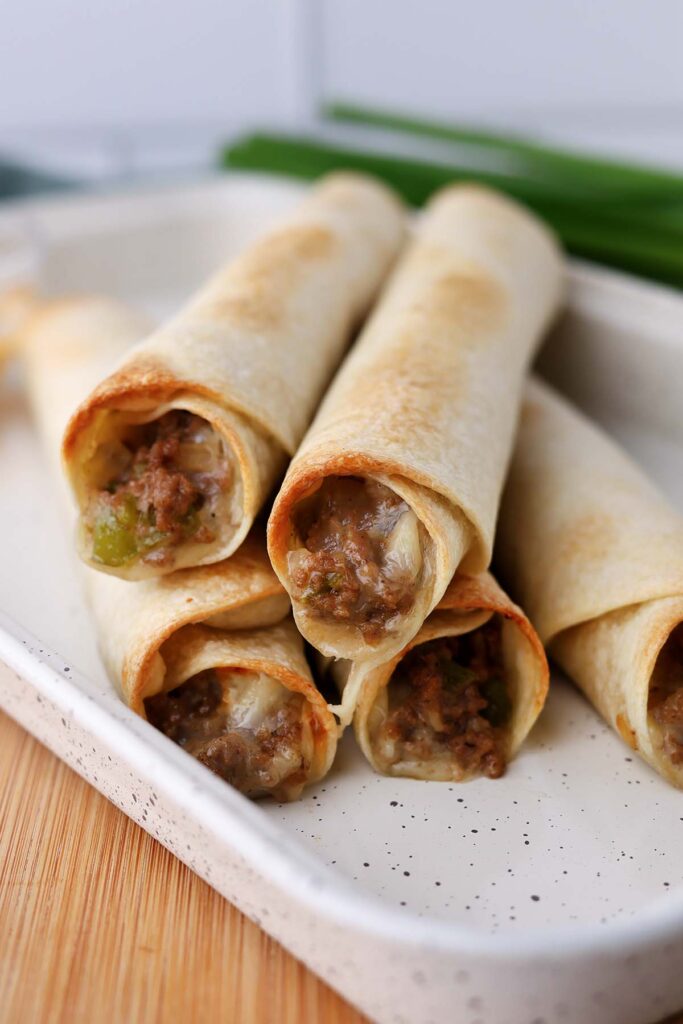close up image showing crispy taquitos on a white tray with cheese in the background
