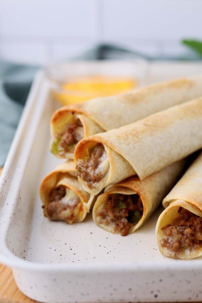 close up image showing crispy taquitos on a white tray with cheese in the background