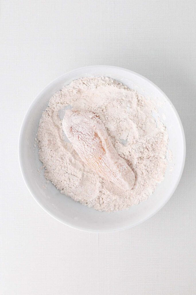 Top down image showing a white bowl filled with flour with one chicken strip inside being coated