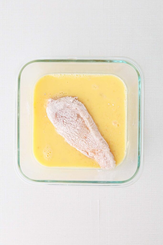 Top down image showing a glass dish with egg whisked inside. A flour coated piece of chicken sits in the middle