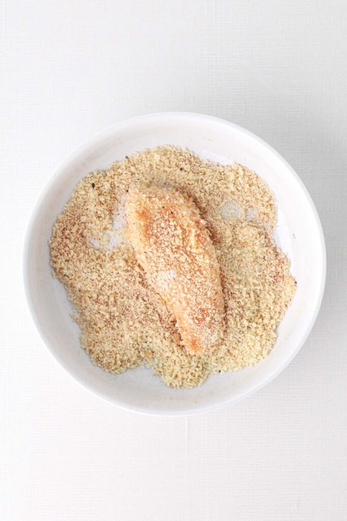 Top down image showing a white bowl filled with bread crumbs with a chicken piece that has been coated in the middle