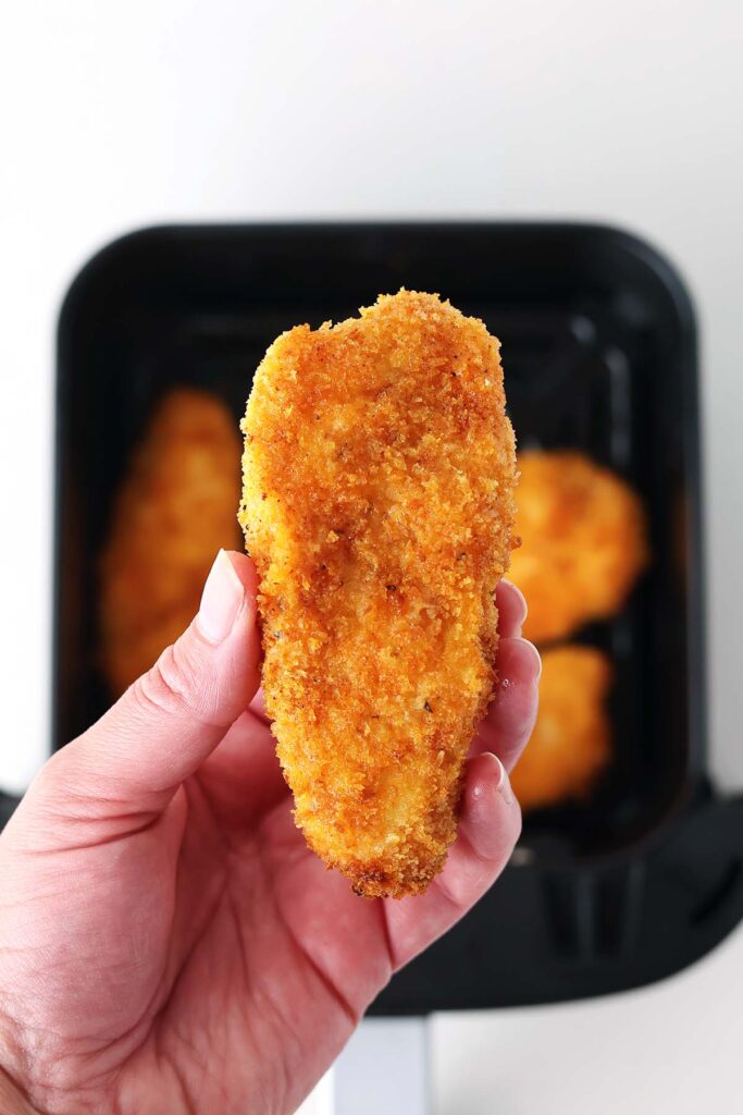 close up image showing a hand holding a crisp chicken piece over the top of an air fryer with more