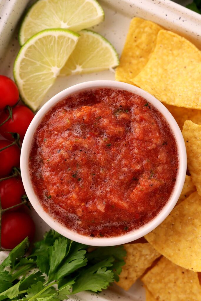 Top down image showing a small white bowl filled with salsa. There are chips, cilantro, tomatoes, and limes on the side.