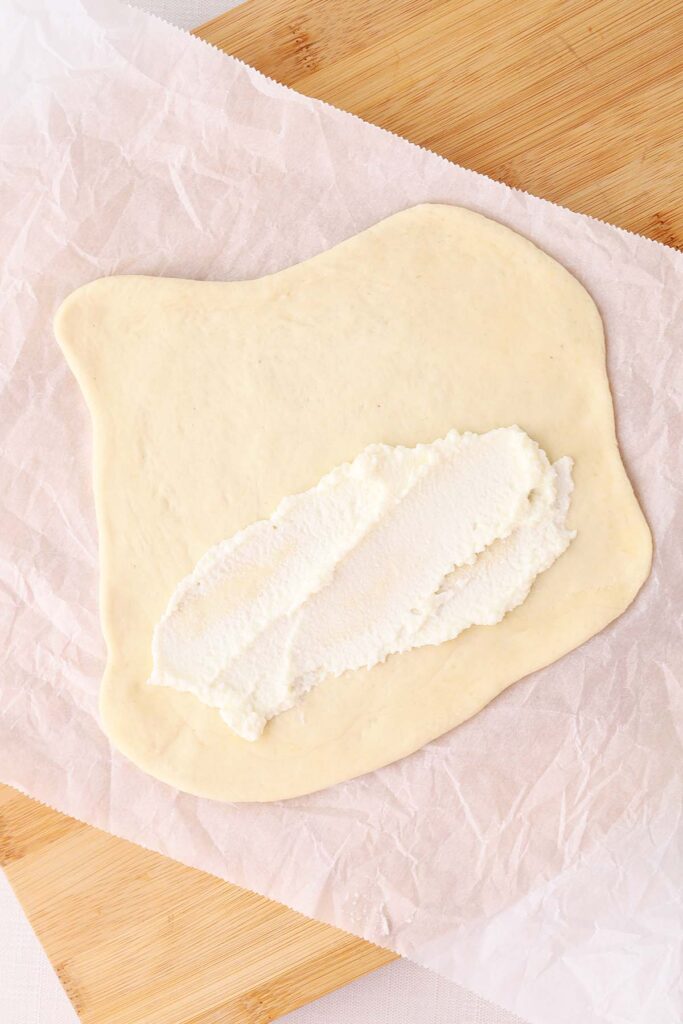 Top down image showing pizza dough rolled out onto a sheet of parchment with ricotta spread across