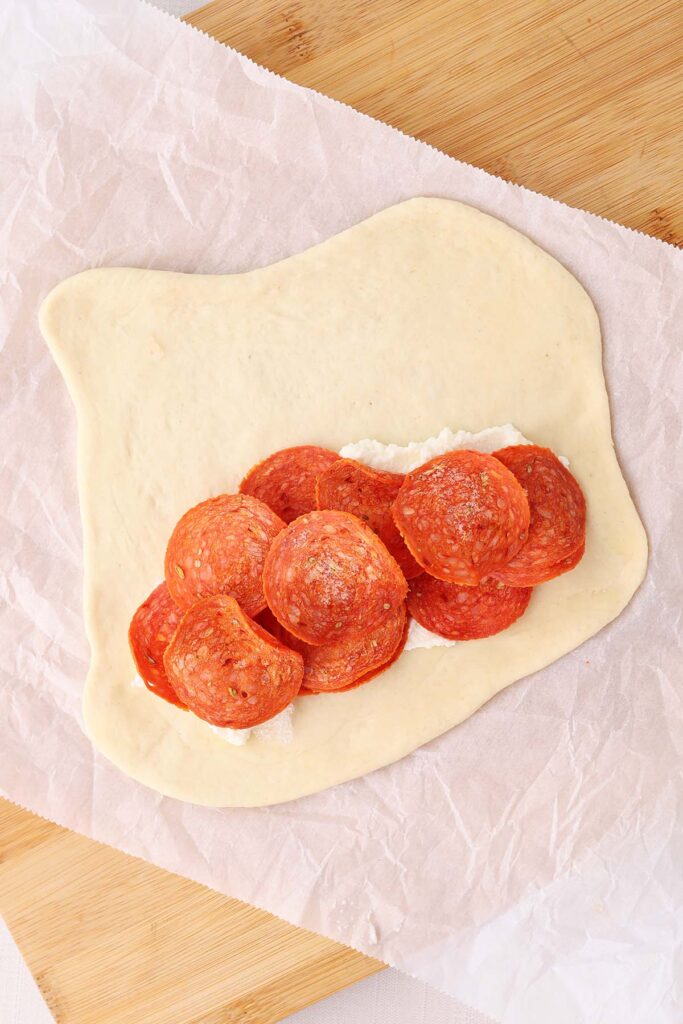 Top down image showing pizza dough rolled out onto a sheet of parchment with ricotta spread across and topped with pepperoni