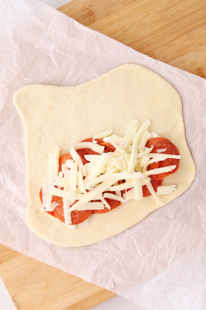 Top down image showing pizza dough rolled out onto a sheet of parchment with ricotta spread across and topped with pepperoni and mozzarella