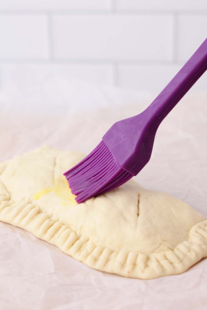 close up image showing a purple pastry brush spreading egg onto the top of an unbaked calzone
