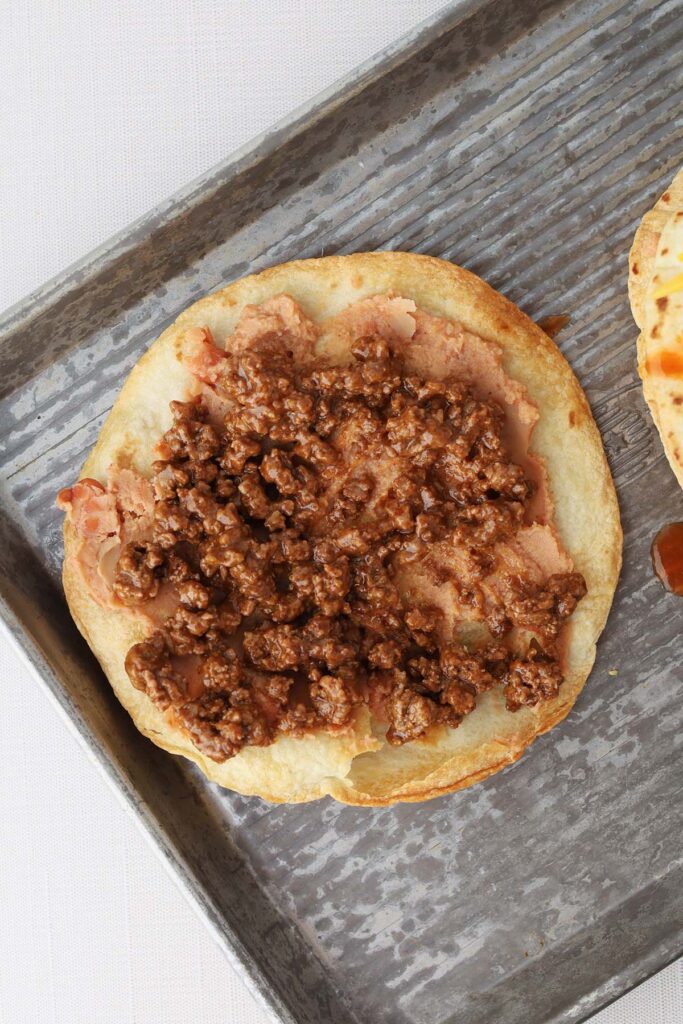 Top down image showing a crispy baked tortilla spread with refried beans and ground beef