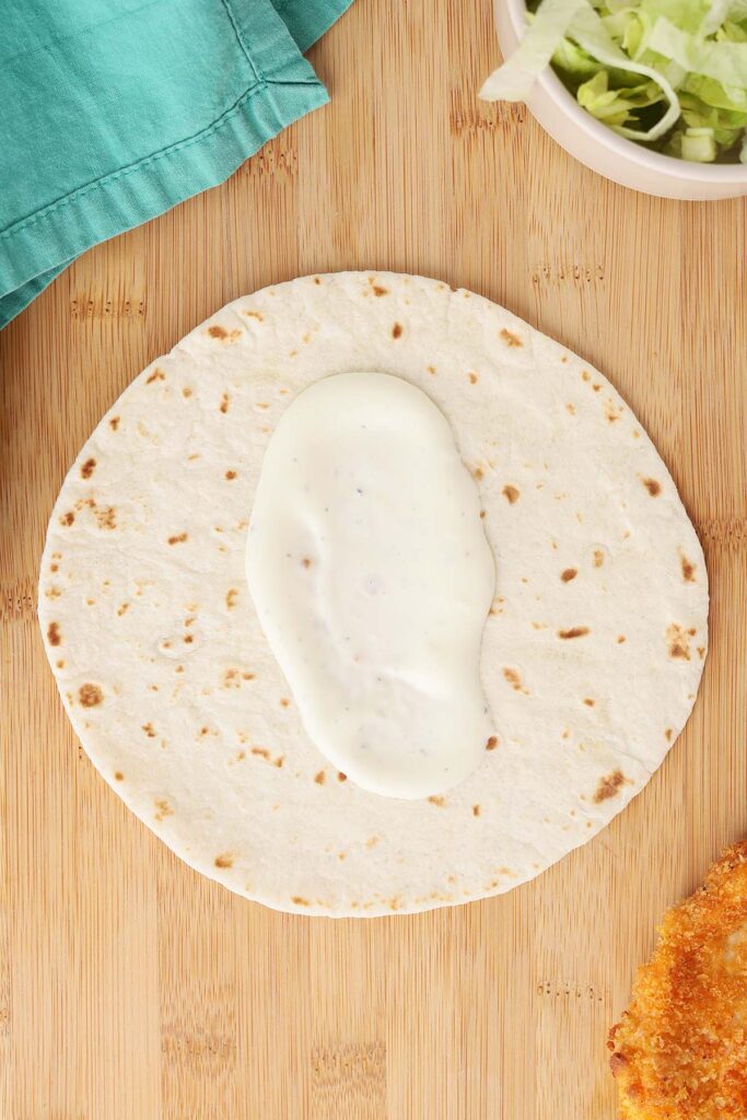 Top down image showing a trotilla on a wooden table top with a spread of ranch dressing. A bowl of lettuch and chicken tender off to the side
