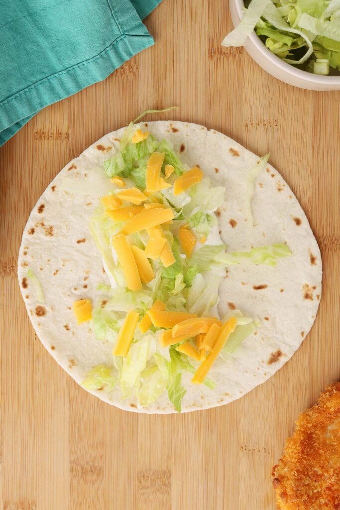 Top down image showing a trotilla on a wooden table top with a spread of ranch dressing and sprinkle of lettuce and cheese. A bowl of lettuch and chicken tender off to the side