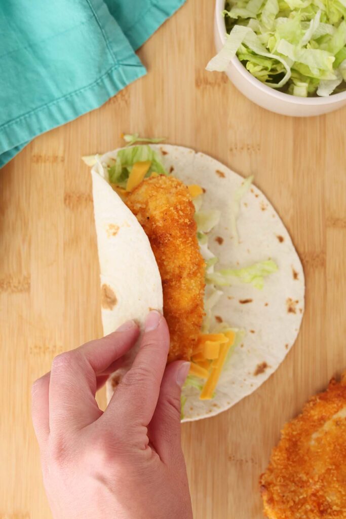 Top down image showing a trotilla on a wooden table top with a spread of ranch dressing and sprinkle of lettuce and cheese with a crispy tender being wrapped. A bowl of lettuch and chicken tender off to the side