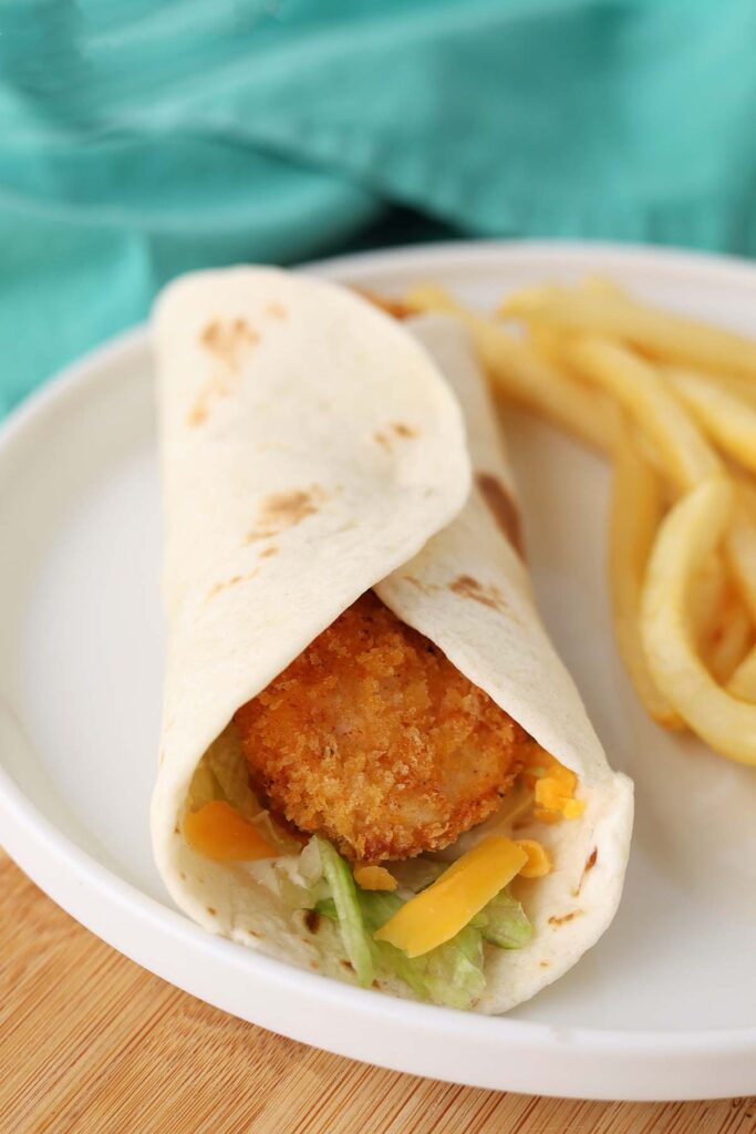 close up image showing a crispy chicken tender wrapped in a tortilla with cheese and lettuce with fries on th e plate to the side