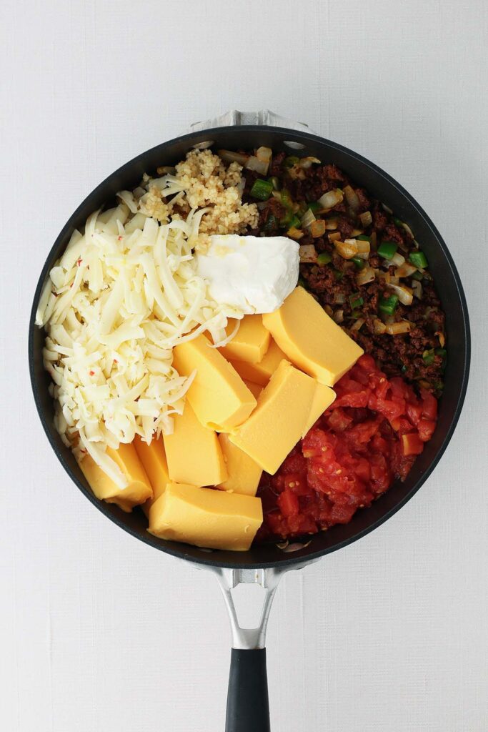 Skillet filled with Velveeta queso ingredients including cubed cheese, shredded pepper jack, cream cheese, cooked sausage, diced tomatoes, onion, jalapeños, and garlic before melting.