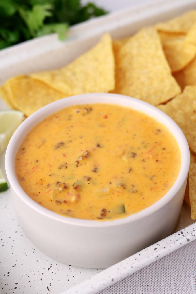 Creamy Velveeta queso dip with sausage and peppers in a white bowl, served with tortilla chips and lime wedges on a tray.