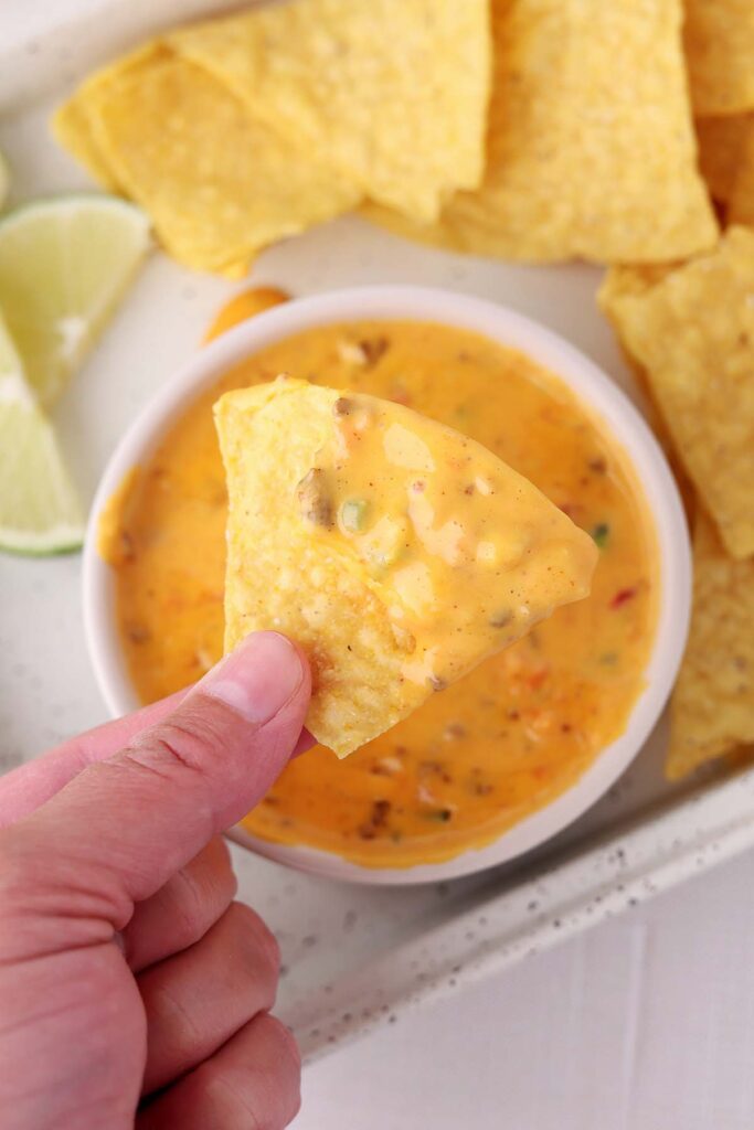 Hand holding a tortilla chip dipped in creamy Velveeta queso with sausage and peppers, served in a white bowl with chips and lime wedges nearby.