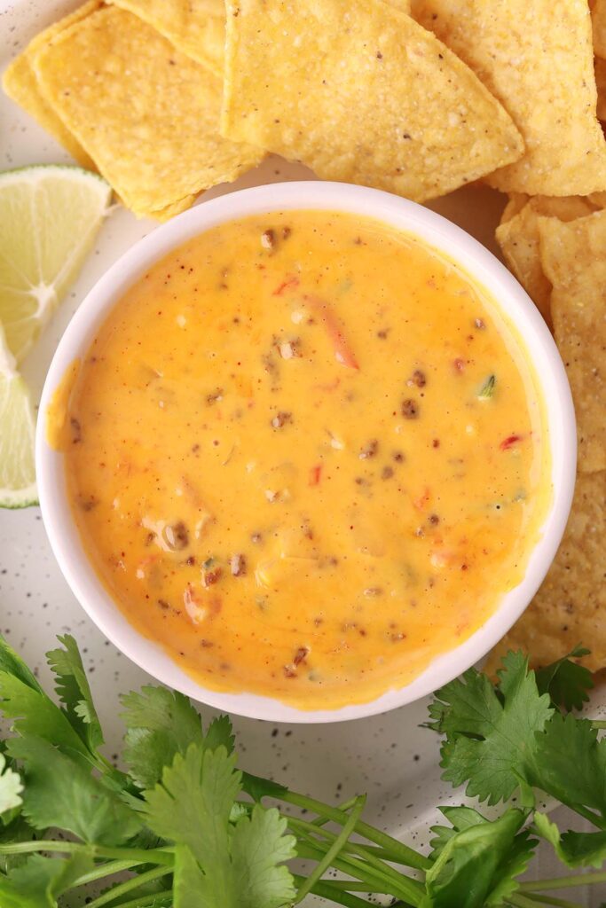Creamy Velveeta queso dip with sausage and peppers in a white bowl, surrounded by tortilla chips, lime wedges, and fresh cilantro.