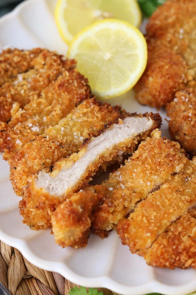 Close up image showing two golden brown chicken cutlets sliced on a white plate with lemon slices