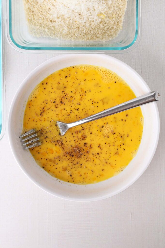 Top down image showing a white round bowl with eggs that have been whisked with a fork with seasonings