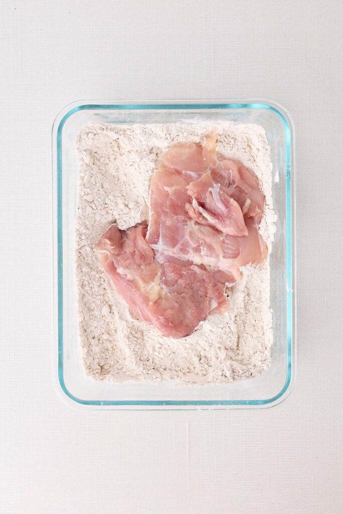 Top down image showing a glass rectangle bowl with flour and chicken tighs
