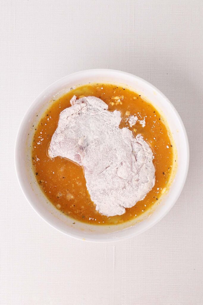 Top down image showing whisked eggs in a round white bowl with chicken that's coated in flour