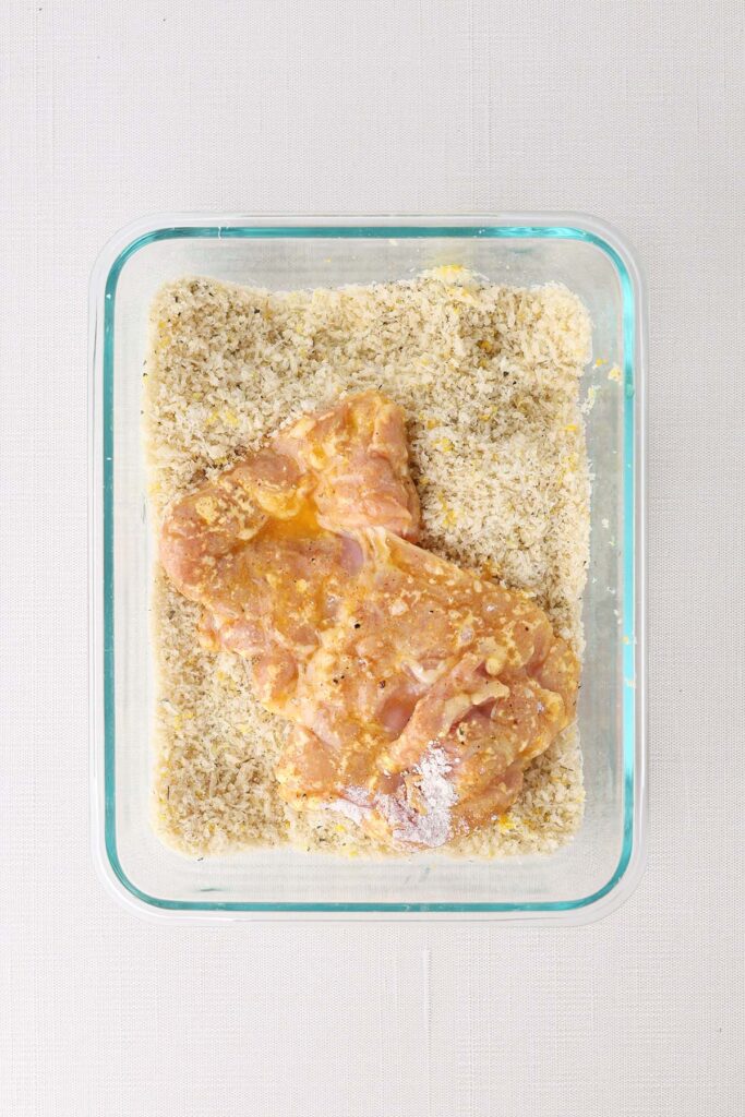 top down image showing a glass rectangle bowl filled with breadcrumbs and a chicken that's coated in flour and egg