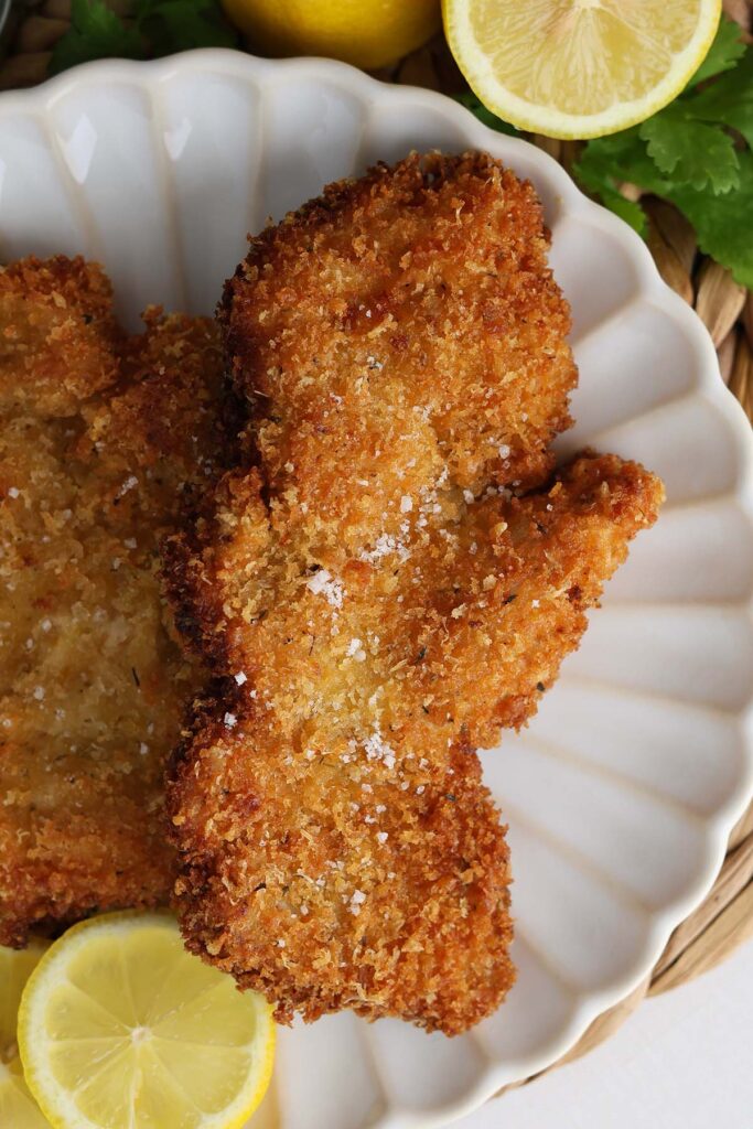 Close up image showing two golden brown chicken cutlets on a white plate with lemon slices