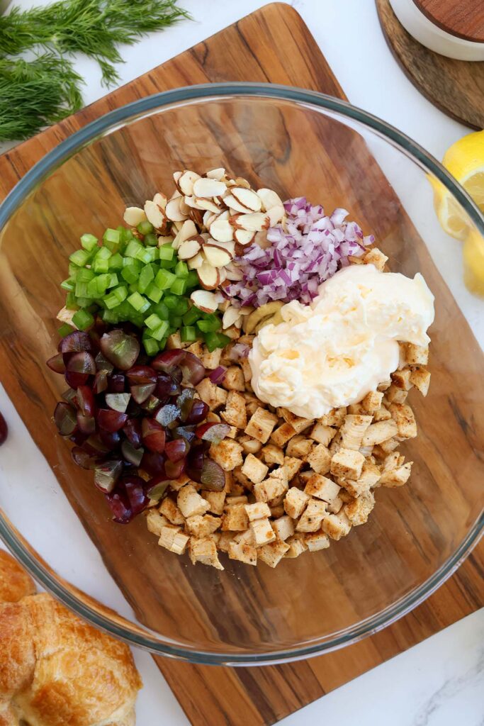 Top down image showing a glass bowl filled with chopped chicken, mayo, chopped grapes, celery, almonds, and red onion unmixed on a wooden cutting board
