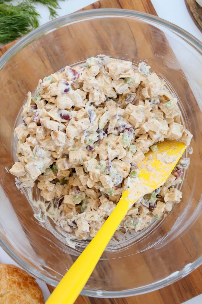 Top down image showing a glass bowl filled with chopped chicken, mayo, chopped grapes, celery, almonds, and red onion mixed on a wooden cutting board