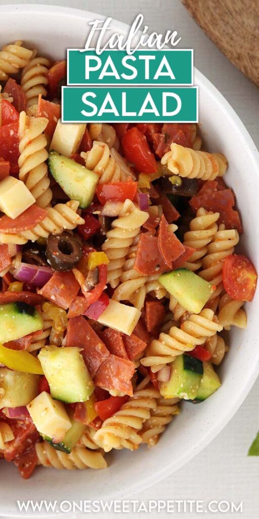 Top down section showing pasta salad in a white dish filled with spiral pasta and chopped veggies. A colored napkin, fresh basil, and a wooden spoon off in the back. Text overlay reads Italian Pasta Salad