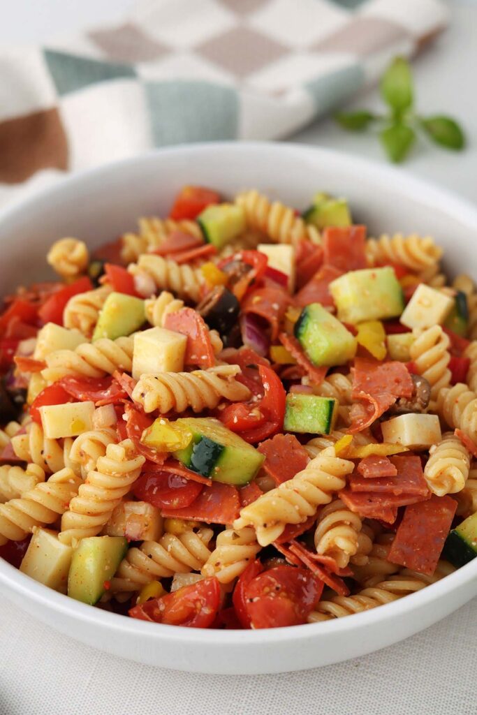 Top down section showing pasta salad in a white dish filled with spiral pasta and chopped veggies. A colored napkin, fresh basil, and a wooden spoon off in the back