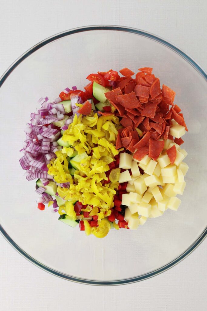 Top down image showing a glass bowl filled with chopped vegetables, cheese, and pepperoni