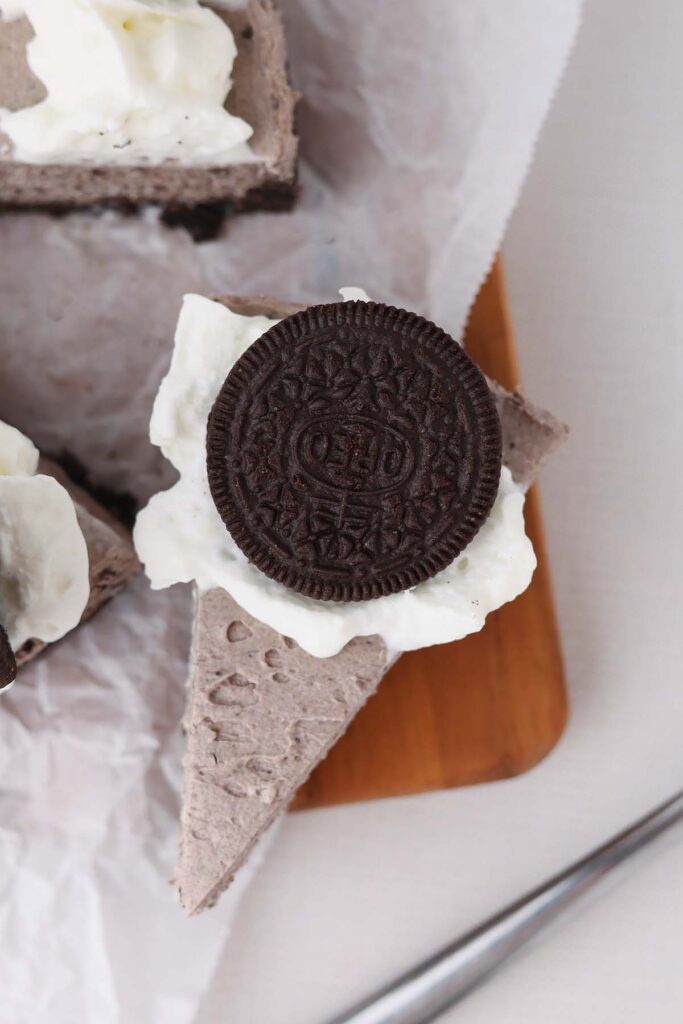 Top down image showing a cookie filled no bake cheesecake slice on a piece of parchment with whipped cream and an oreo on top
