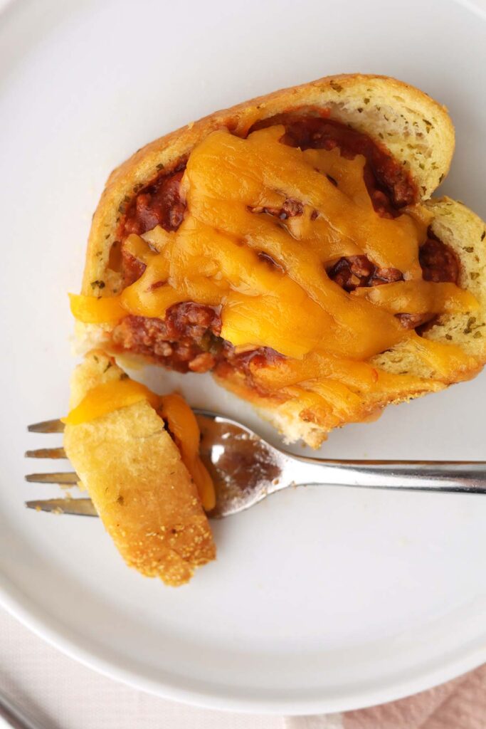 Close up image showing a slice of garlic toast topped with a sauce beef mix and melted cheese on a white plate. One bite is sitting on a fork.