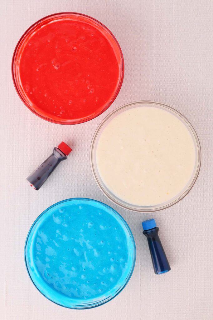 Top down image showing three glass bowls each filled with cupcake batter. Red, white and blue. there are also two food color bottles next to the bowls in red and blue colors