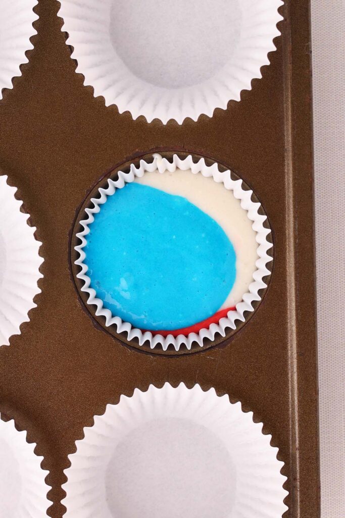 Top down image showing a closeup of a cupcake tin with white paper liners. One liner has red white and blue cake batter inside