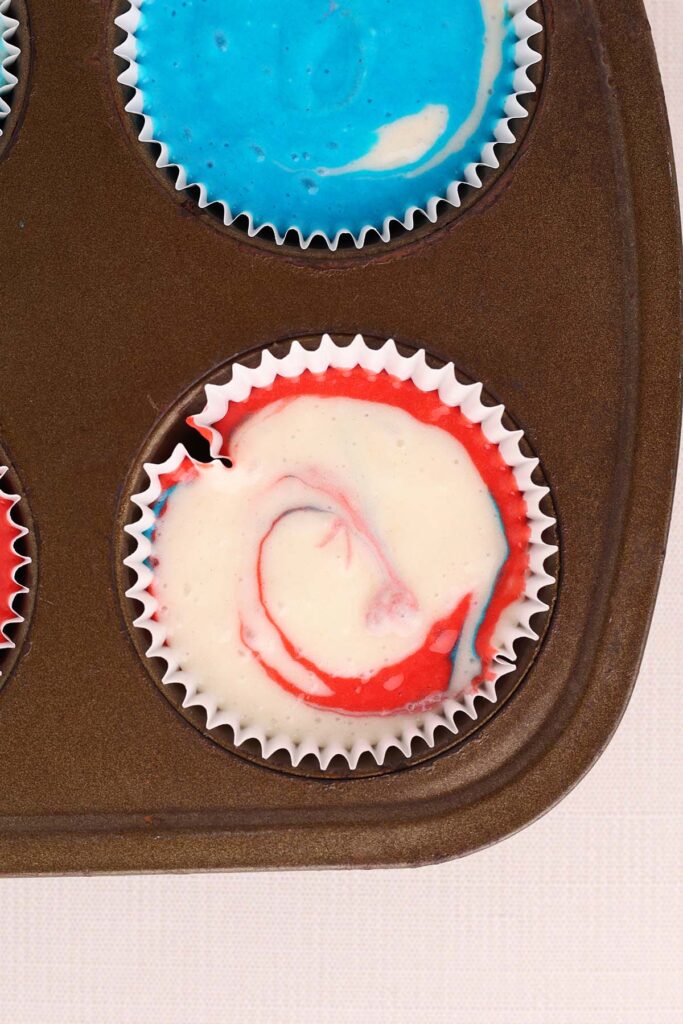 Top down image showing a closeup of a cupcake tin with white paper liners. One liner has red white and blue cake batter inside swirled