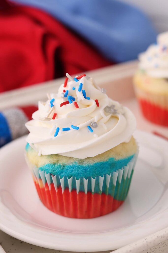 Close up image showing a red white and blue cupcake with white frosting and festive sprinkles on a white plate.