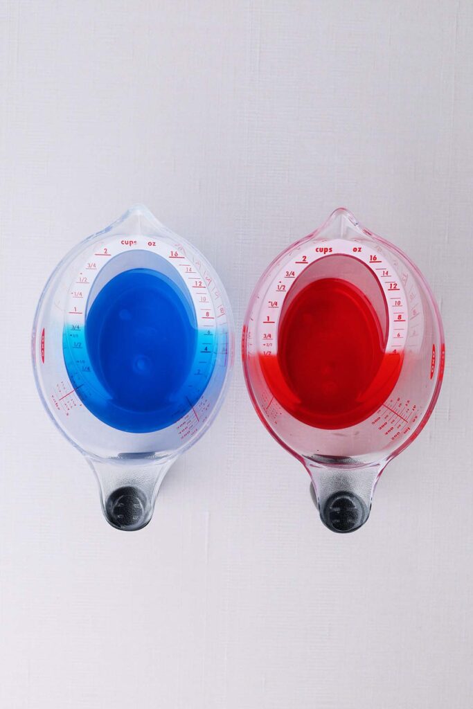 Top down image showing two liquid measuring cups. One filled with blue liquid one filled with red.