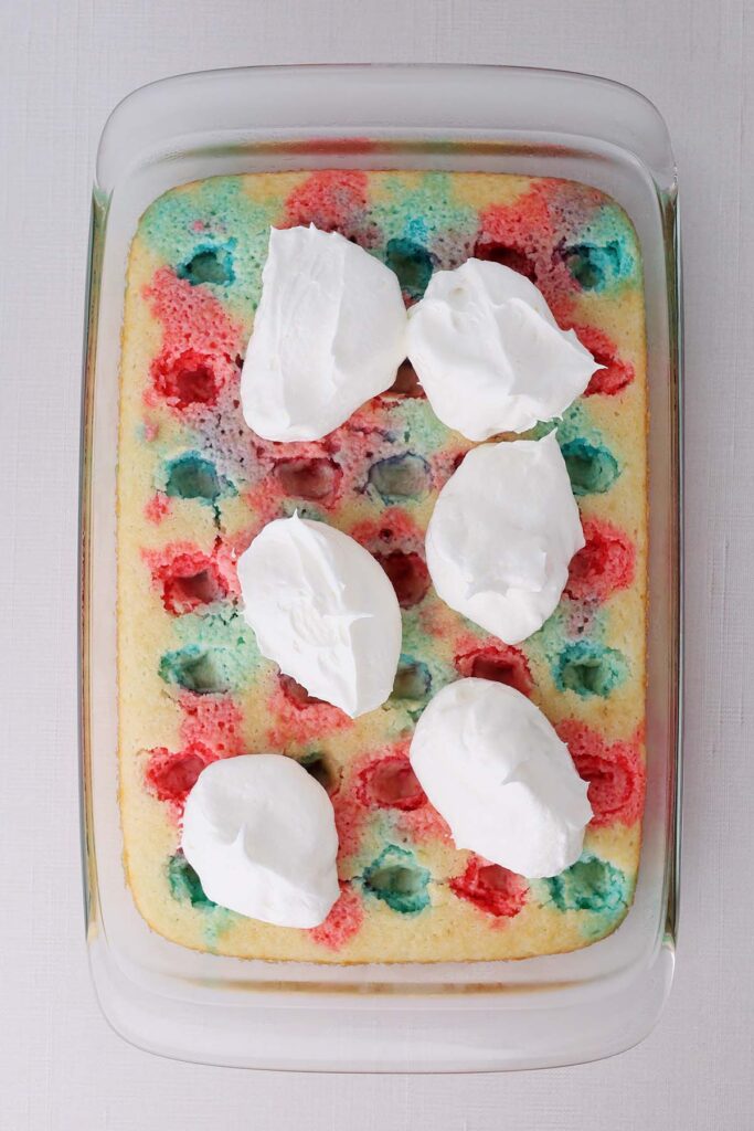 top down image showing a glass pan with a cake that has holes poked in it and red and blue liquid alternating each hole. Dollops of whipped topping on top