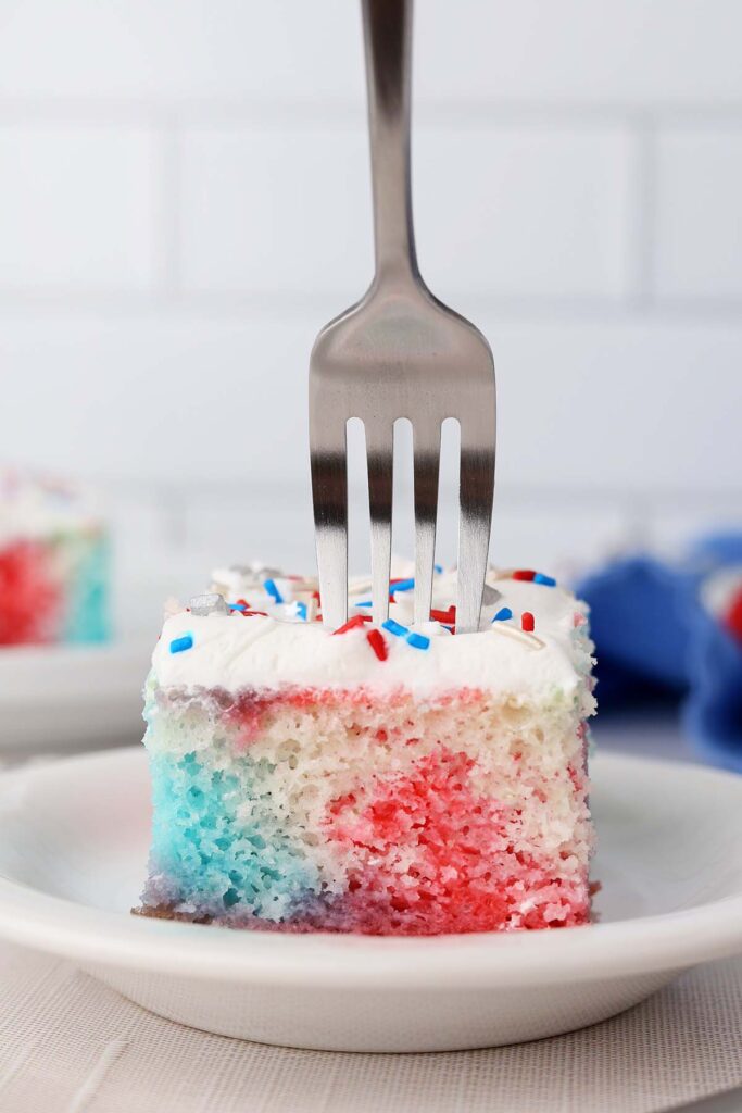 close up image showing a slice of red white and blue cake with white frosting and red white and blue sprinkles. A fork is being pushed into the top