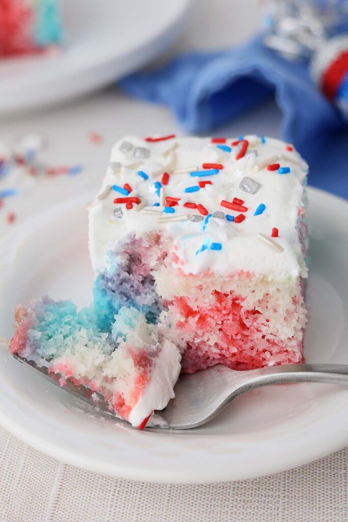 Close up image showing a red white and blue colored cake with white topping and sprinkles. A bite is sitting on a fork