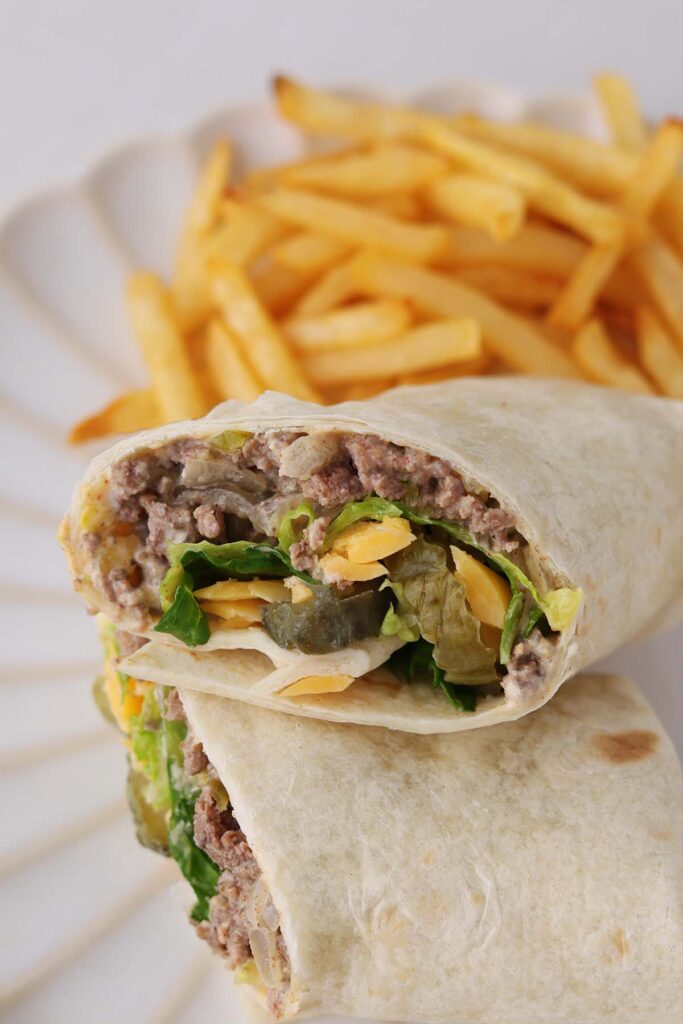 Close up image showing a tortilla filled with ground beef, cheese, lettuce, and pickles stacked on a plate with fries