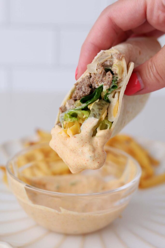 Close up image showing a tortilla filled with ground beef, cheese, lettuce, and pickles dipped in fry sauce