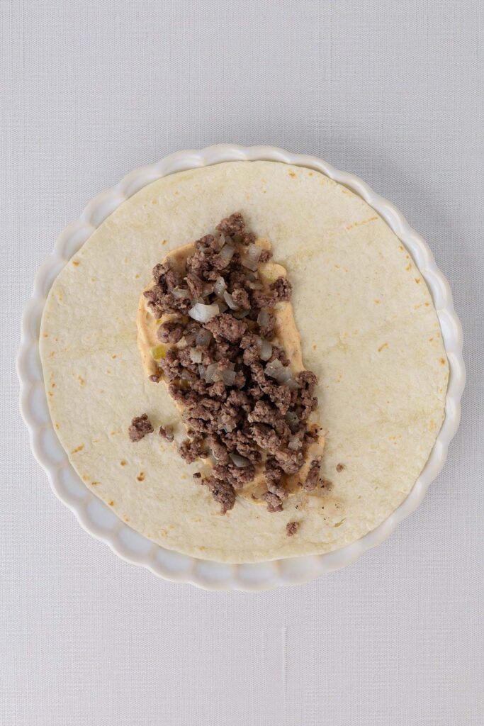 Top down image showing a tortilla on a white plate with burger sauce spread and topped with cooked beef